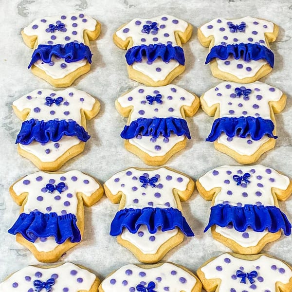 Rows of baby onesie-shaped cookies with white icing, purple polka dots, and blue ruffled frosting.
