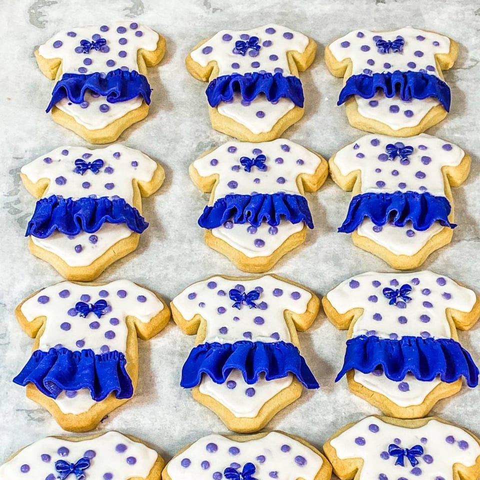 Rows of baby onesie-shaped cookies with white icing, purple polka dots, and blue ruffled frosting.