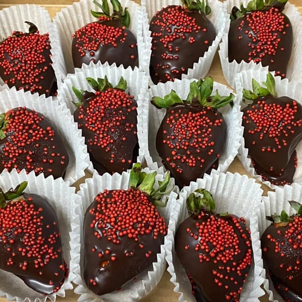 A dozen chocolate-covered strawberries with red sprinkles, each in a white paper cup.