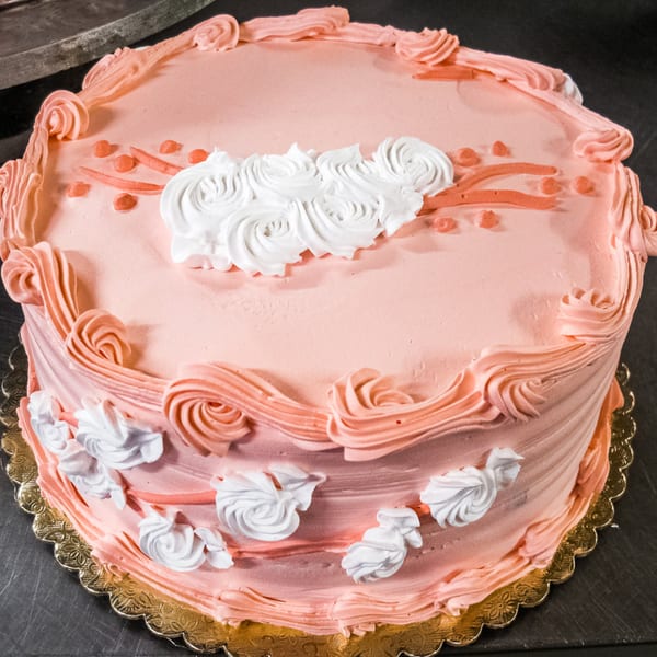 A pink frosted cake with white and pink icing swirls and small heart decorations from L.A. Bakery.