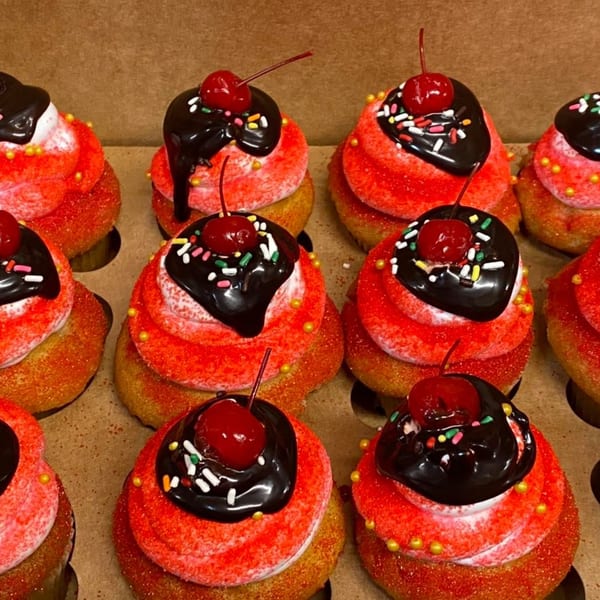 Rows of L.A. Bakery cupcakes with red frosting, chocolate ganache, sprinkles, and a cherry on top.